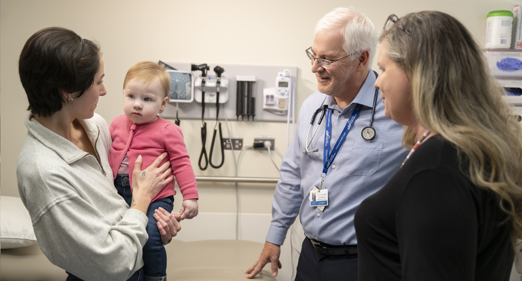Dr. Peter Blaikie in his examination room talking with a nurese and a patient who is holding a baby.